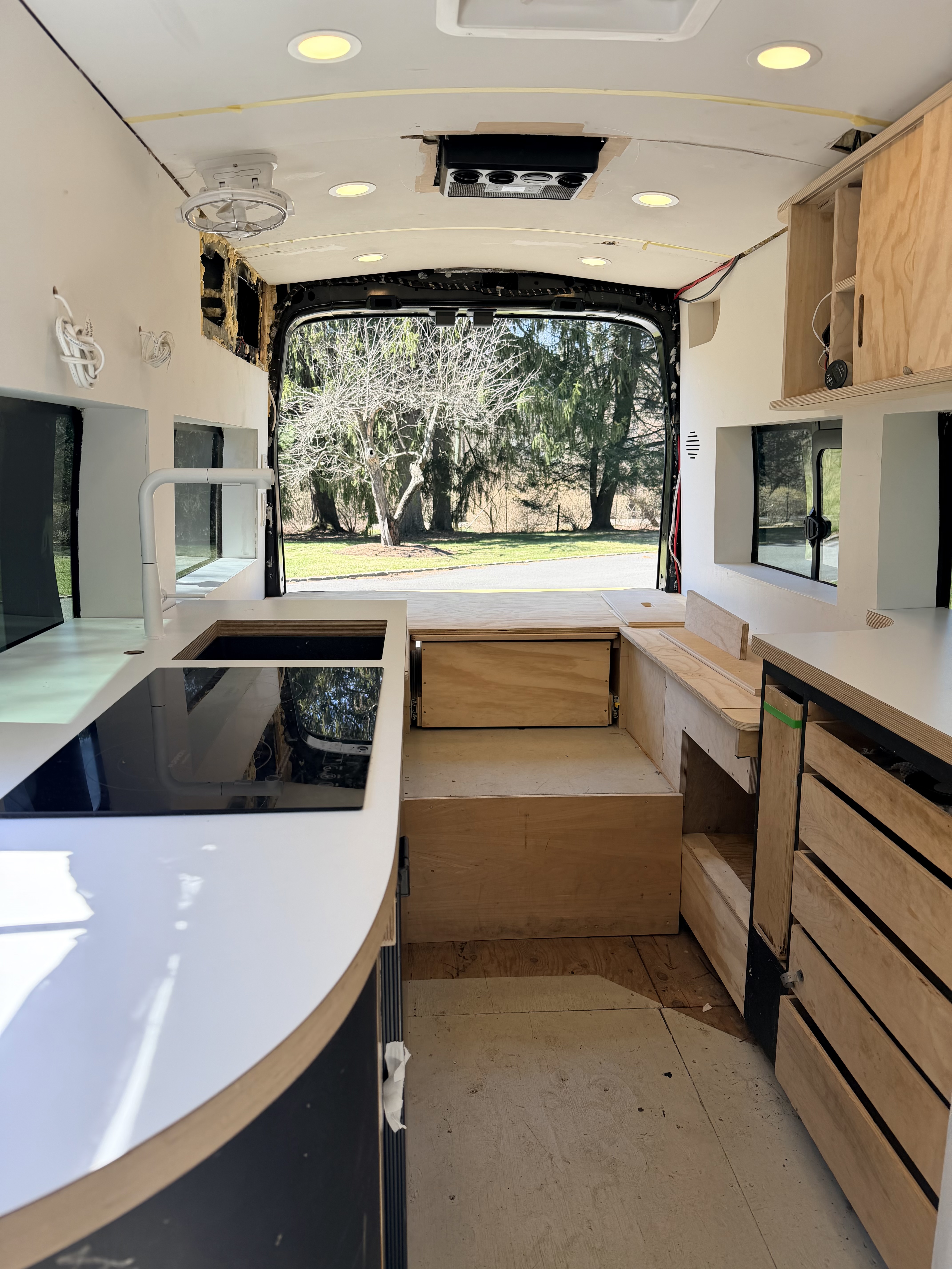 Actual unfinished van interior with counters and cooktop in place, exposed cabinet openings, bare floor and unfinished ceiling edges